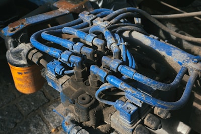 A complex network of blue hydraulic tubes connected to a metal machinery component, with a visible orange filter bearing text in the background. The surface exhibits signs of wear and dirt, indicating usage and exposure to industrial conditions.