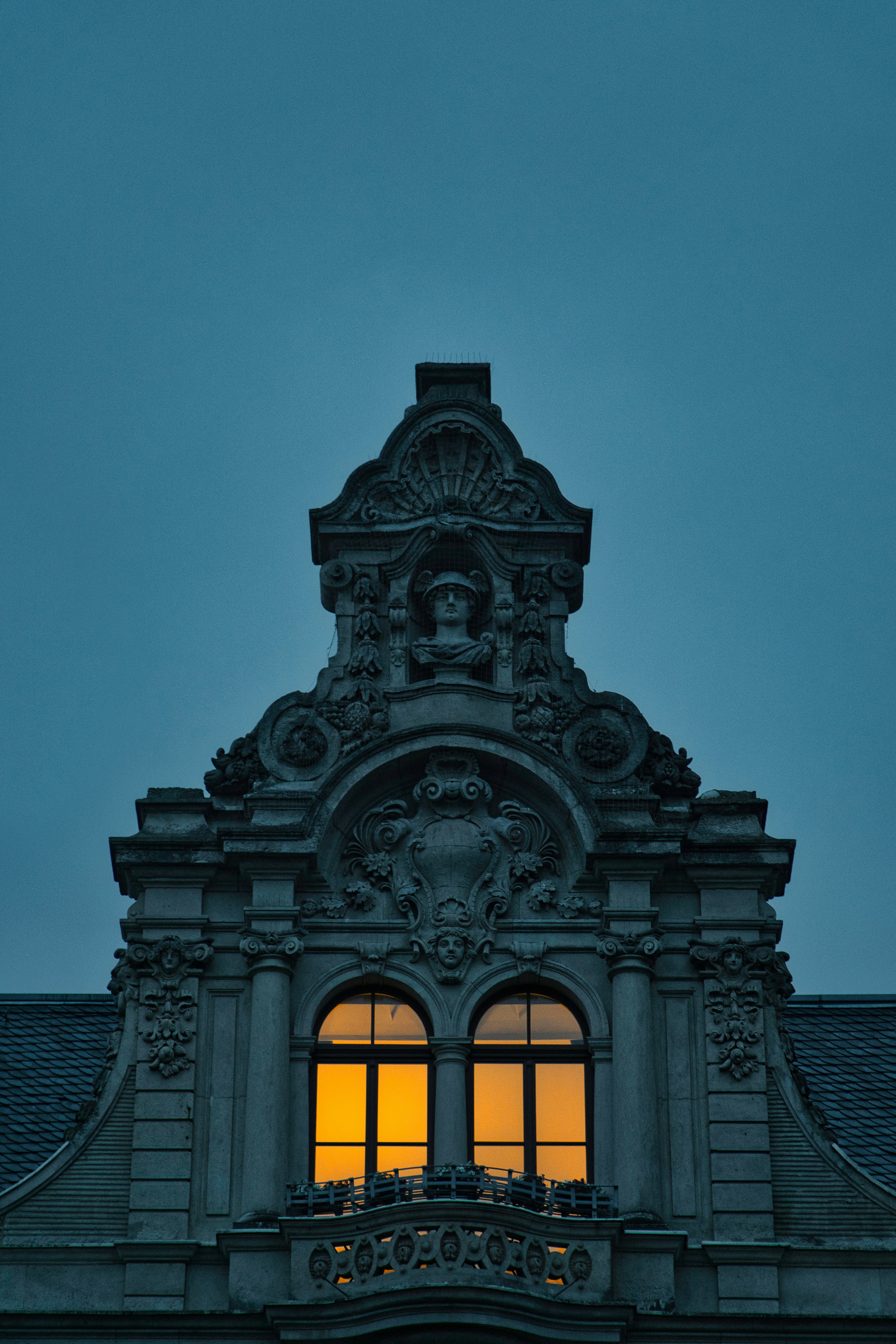 Dramatic architectural exterior at dusk, deep shadows, warm amber interior glow visible through floor-to-ceiling windows, dark stone facade