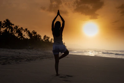 Sunset silhouette of a yoga practitioner balancing in tree pose on the beach.