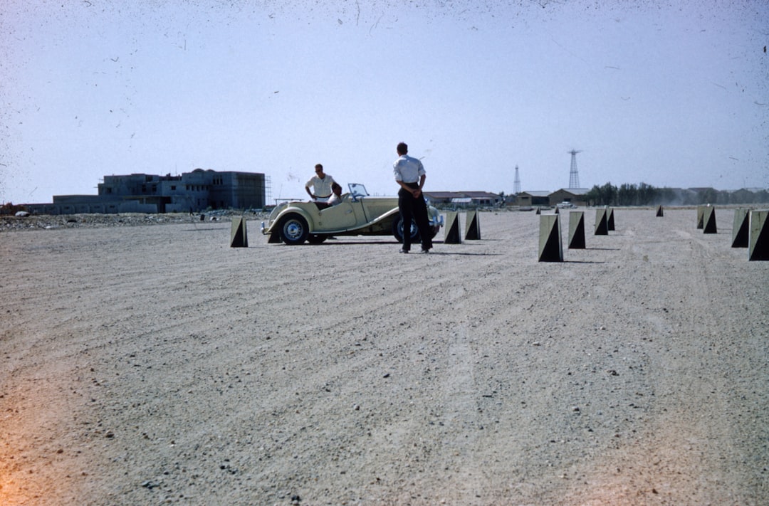 man in black jacket and black pants sitting on white car on gray sand during daytime