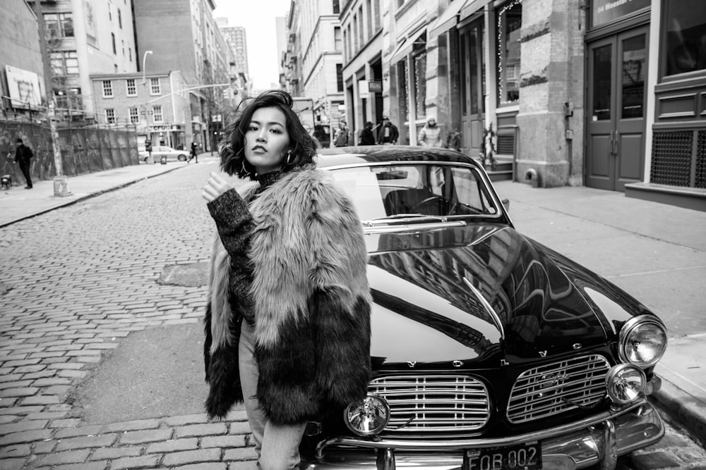 grayscale photo of woman in fur coat standing on sidewalk