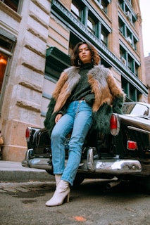 A person is leaning against the back of a black vintage car, wearing a stylish multicolored fur coat, dark sweater, blue jeans, and white heeled boots. The urban backdrop features a classic building with large windows.