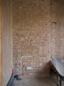 A partially constructed or renovated brick wall displaying a rough, textured surface. There is a notable arch shape embedded within the wall, which is not fully defined. Below the arch, an electrical outlet is installed with cables running vertically and horizontally, partially plastered over. Dust and debris cover the floor, and there is a hint of a wheelbarrow to the right side of the image.