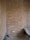 A partially constructed or renovated brick wall displaying a rough, textured surface. There is a notable arch shape embedded within the wall, which is not fully defined. Below the arch, an electrical outlet is installed with cables running vertically and horizontally, partially plastered over. Dust and debris cover the floor, and there is a hint of a wheelbarrow to the right side of the image.