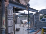 A construction site featuring partially constructed walls made of bricks and steel beams. Scaffolding and construction tools are present, along with partially erected structures. The background includes a garden area with some trees and a cloudy sky.
