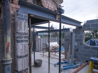 A construction site featuring partially constructed walls made of bricks and steel beams. Scaffolding and construction tools are present, along with partially erected structures. The background includes a garden area with some trees and a cloudy sky.