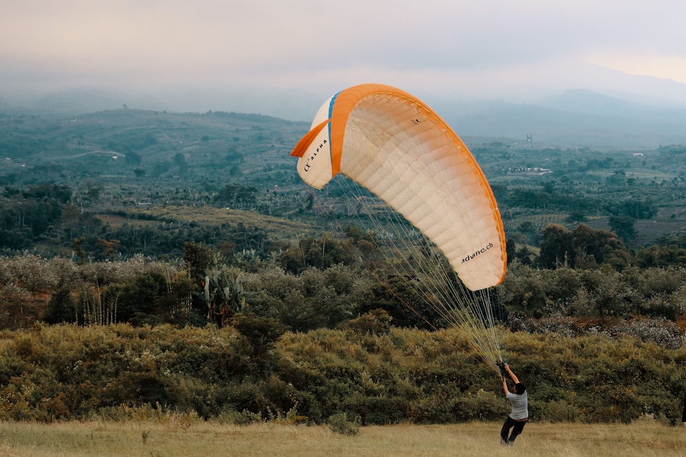 Person In Black Jacket And Blue Denim Jeans Riding Orange Parachute Over Green Mountains During Daytime