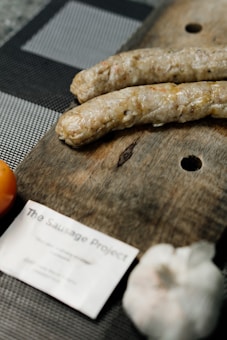 Two sausages rest on a wooden cutting board, with a blurred note labeled 'The Sausage Project' nearby. Partially visible are a tomato and a bulb of garlic surrounding the board. The surface beneath appears to be a gray checkered fabric.