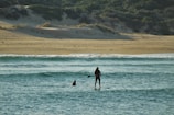Two people are engaging in water activities near the shore. One person is on a paddleboard, standing and using a paddle, while the other is in the water nearby. The backdrop includes a sandy beach and lush, green vegetation on dunes.