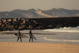 Two surfers walk along a sandy beach, each holding a surfboard. They are wearing wetsuits and heading towards a section of the beach where waves break gently. A rocky breakwater or jetty is visible in the background, with mountains and sand dunes on the horizon under a partly cloudy sky.