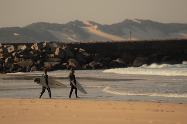 Two surfers walk along a sandy beach, each holding a surfboard. They are wearing wetsuits and heading towards a section of the beach where waves break gently. A rocky breakwater or jetty is visible in the background, with mountains and sand dunes on the horizon under a partly cloudy sky.