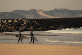 Two surfers walk along a sandy beach, each holding a surfboard. They are wearing wetsuits and heading towards a section of the beach where waves break gently. A rocky breakwater or jetty is visible in the background, with mountains and sand dunes on the horizon under a partly cloudy sky.