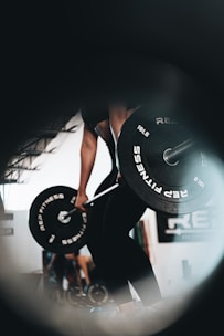 A person is performing a deadlift with a barbell in a gym. The perspective is low and centered, capturing the weights prominently. The environment suggests a focused and determined atmosphere.