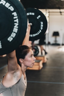 A woman is lifting a barbell overhead in a gym setting. Her expression is focused and determined as she performs the exercise. The gym environment includes equipment like benches and weights in the background, with a slightly blurred effect, emphasizing the subject.