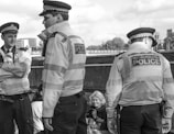 Three police officers in uniforms, wearing hats and jackets with 'Metropolitan Police' labels, stand near a railing. They appear to be conversing or looking at something outside the frame. Nearby, an older woman is seated, looking down with a slight smile. The scene appears to be in an urban area with buildings and trees in the background.