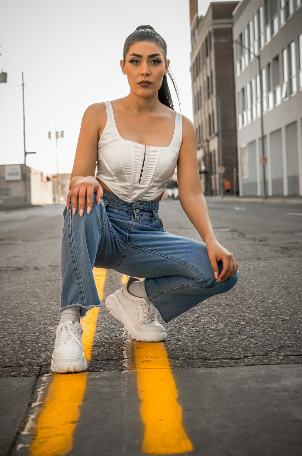 Mujer con camiseta blanca sin mangas y jeans de mezclilla azules sentada en un camino de concreto gris durante el día