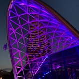 A futuristic cityscape featuring a geometric metal framework illuminated with vibrant purple and blue lights. In the background, a modern skyscraper is lit with horizontal red lines, set against a twilight sky.