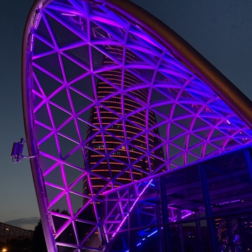 A futuristic cityscape featuring a geometric metal framework illuminated with vibrant purple and blue lights. In the background, a modern skyscraper is lit with horizontal red lines, set against a twilight sky.