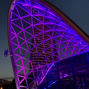 A futuristic cityscape featuring a geometric metal framework illuminated with vibrant purple and blue lights. In the background, a modern skyscraper is lit with horizontal red lines, set against a twilight sky.