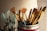 A collection of paintbrushes with golden bristles and handles is arranged in a container. The scene is warmly lit, highlighting the textures of the bristles. In the background, several light blue containers, possibly paint supplies, are slightly out of focus.