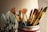 A collection of paintbrushes with golden bristles and handles is arranged in a container. The scene is warmly lit, highlighting the textures of the bristles. In the background, several light blue containers, possibly paint supplies, are slightly out of focus.