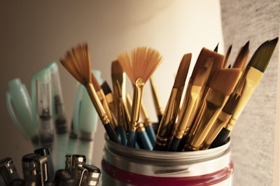 A collection of paintbrushes with golden bristles and handles is arranged in a container. The scene is warmly lit, highlighting the textures of the bristles. In the background, several light blue containers, possibly paint supplies, are slightly out of focus.