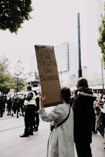 A person is holding a sign that reads 'Teach the truth in school' in the midst of a crowd. The scene appears to be a protest or rally, with people gathered around and some wearing security uniforms. The surroundings suggest an urban setting with buildings visible in the background.