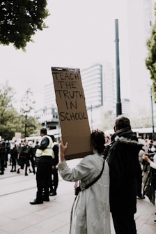 A person is holding a sign that reads 'Teach the truth in school' in the midst of a crowd. The scene appears to be a protest or rally, with people gathered around and some wearing security uniforms. The surroundings suggest an urban setting with buildings visible in the background.