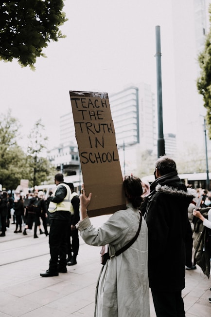A person is holding a sign that reads 'Teach the truth in school' in the midst of a crowd. The scene appears to be a protest or rally, with people gathered around and some wearing security uniforms. The surroundings suggest an urban setting with buildings visible in the background.