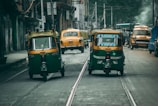 Two auto rickshaws drive on a city street lined with buildings. Several yellow taxis, including one parked on the side, are visible in the background. A bus and a few pedestrians can also be seen. The road appears used, as demonstrated by the presence of tram tracks.