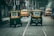 Two auto rickshaws drive on a city street lined with buildings. Several yellow taxis, including one parked on the side, are visible in the background. A bus and a few pedestrians can also be seen. The road appears used, as demonstrated by the presence of tram tracks.