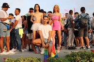 A group of people are gathered outdoors, some wearing colorful clothing and pride flags. In the center foreground, three individuals pose confidently. The crowd in the background is engaged in conversation and appears festive, with pink inflatable decorations visible.