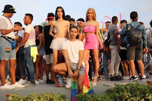 A group of people are gathered outdoors, some wearing colorful clothing and pride flags. In the center foreground, three individuals pose confidently. The crowd in the background is engaged in conversation and appears festive, with pink inflatable decorations visible.