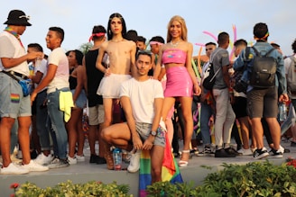 A group of people are gathered outdoors, some wearing colorful clothing and pride flags. In the center foreground, three individuals pose confidently. The crowd in the background is engaged in conversation and appears festive, with pink inflatable decorations visible.
