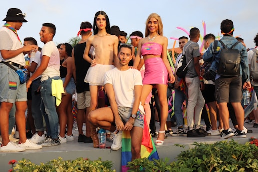 A group of people are gathered outdoors, some wearing colorful clothing and pride flags. In the center foreground, three individuals pose confidently. The crowd in the background is engaged in conversation and appears festive, with pink inflatable decorations visible.