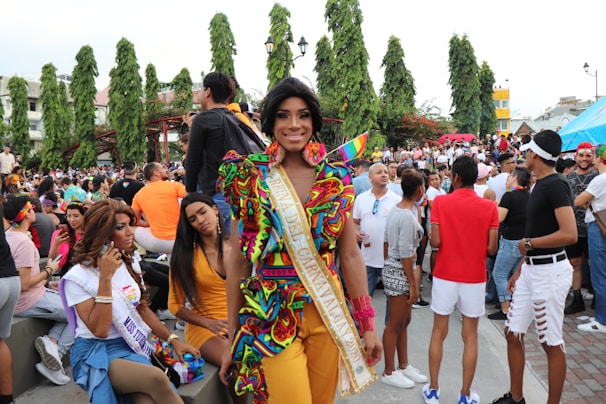 A vibrant crowd gathers outdoors, with a focus on an individual wearing a colorful, patterned outfit and a sash. The background includes people interacting, some on phones, others standing or sitting in various casual attire. Lush green trees and urban structures form the backdrop, creating a lively atmosphere.