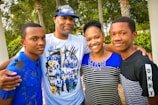 A family of four stands closely together outdoors, smiling warmly. The group includes two adults and two young individuals, all dressed in casual clothing with a backdrop of trees and greenery.