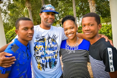 A family of four stands closely together outdoors, smiling warmly. The group includes two adults and two young individuals, all dressed in casual clothing with a backdrop of trees and greenery.