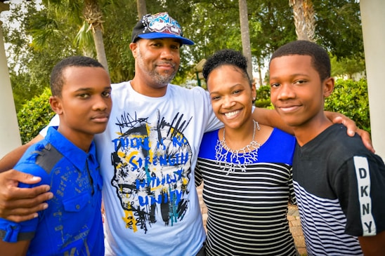 A family of four stands closely together outdoors, smiling warmly. The group includes two adults and two young individuals, all dressed in casual clothing with a backdrop of trees and greenery.