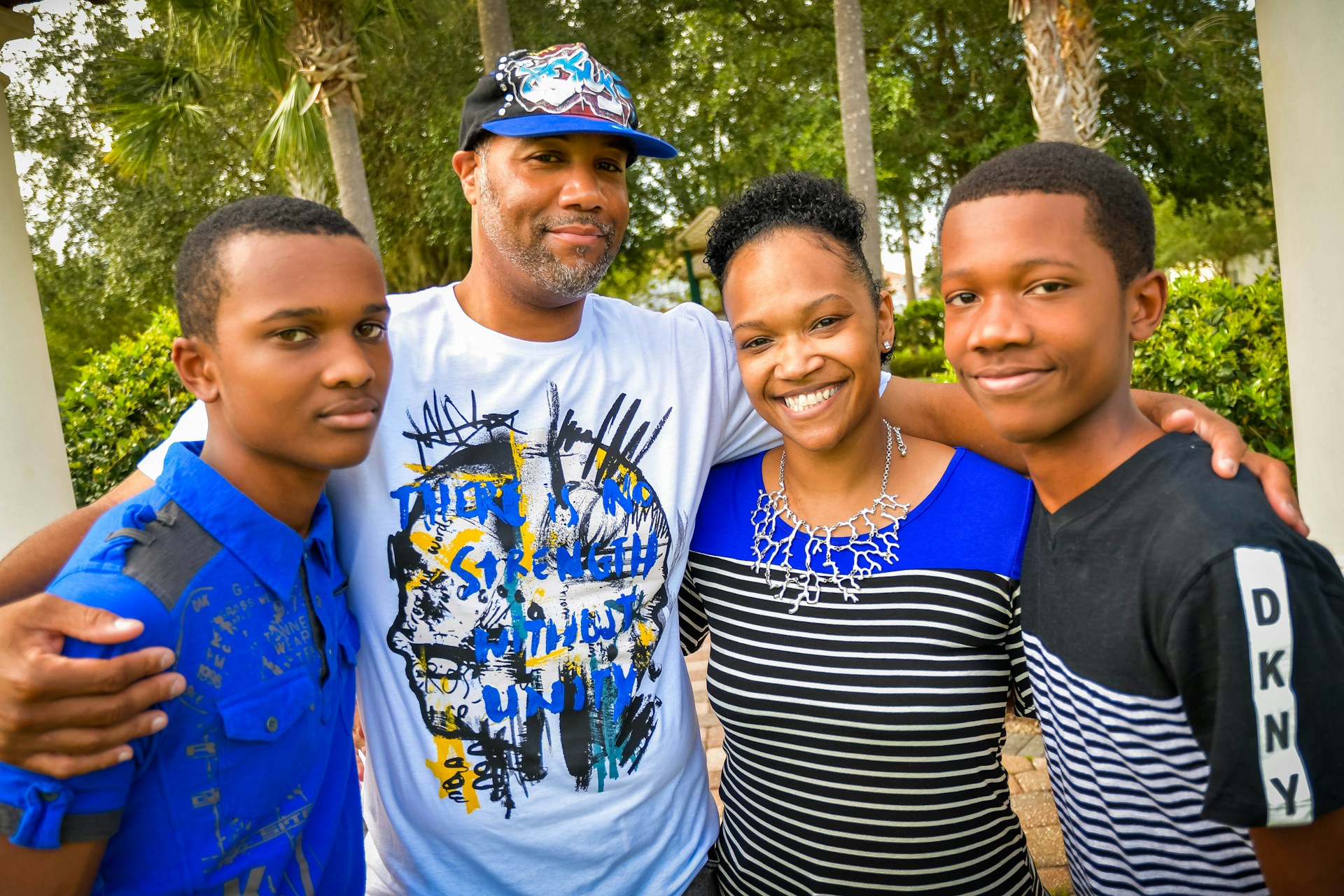 A family of four stands closely together outdoors, smiling warmly. The group includes two adults and two young individuals, all dressed in casual clothing with a backdrop of trees and greenery.