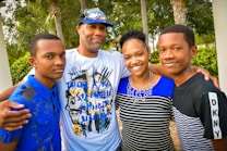 A family of four stands closely together outdoors, smiling warmly. The group includes two adults and two young individuals, all dressed in casual clothing with a backdrop of trees and greenery.