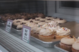 Rows of cupcakes displayed in a bakery case, topped with swirls of creamy frosting and colorful sprinkles. The front row showcases vanilla cupcakes labeled 'Villy Nilly', while other varieties are visible in the background.
