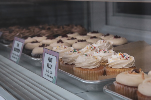 Rows of colorful cupcakes with creamy frosting and sprinkles displayed on a bakery shelf.