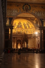 A grand cathedral interior with intricate mosaics depicting religious figures. The warm lighting highlights the golden hues of the artwork, creating a solemn and reverent atmosphere. People are scattered throughout the spacious hall, some gathered in groups, admiring the architecture.