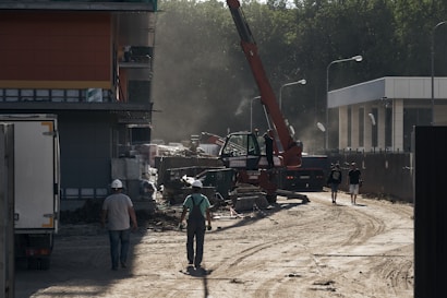 A construction site featuring a large crane in operation surrounded by various building materials. Several individuals wearing safety helmets are walking in different directions, implying a busy work environment. The setting has dusty air with sunlight filtering through, casting shadows and enhancing the industrial atmosphere.