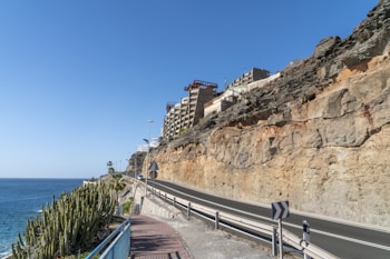 A coastal road with a rocky cliff on the right and the ocean on the left. Several apartment buildings are situated on top of the cliff. The road is lined with a metal guardrail, and there's a walkway with cacti planted along it. The sky is clear and blue.