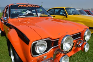 A vintage orange Ford car with the word 'MEXICO' on the windshield is parked on a grassy area. The car is equipped with additional spotlights on the front bumper. Several other colorful classic cars are lined up in the background, including a yellow one.