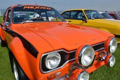 A vintage orange Ford car with the word 'MEXICO' on the windshield is parked on a grassy area. The car is equipped with additional spotlights on the front bumper. Several other colorful classic cars are lined up in the background, including a yellow one.