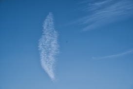 A wisp of white cloud stretches vertically against a bright blue sky, forming a feather-like shape. Thin, wispy clouds are scattered to the right, creating a serene backdrop.