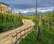 A scenic rural pathway winds through a lush green vineyard, flanked by wooden posts and a rustic fence. In the background, snow-capped mountains rise majestically under a slightly overcast sky, adding a dramatic contrast to the vibrant greenery.